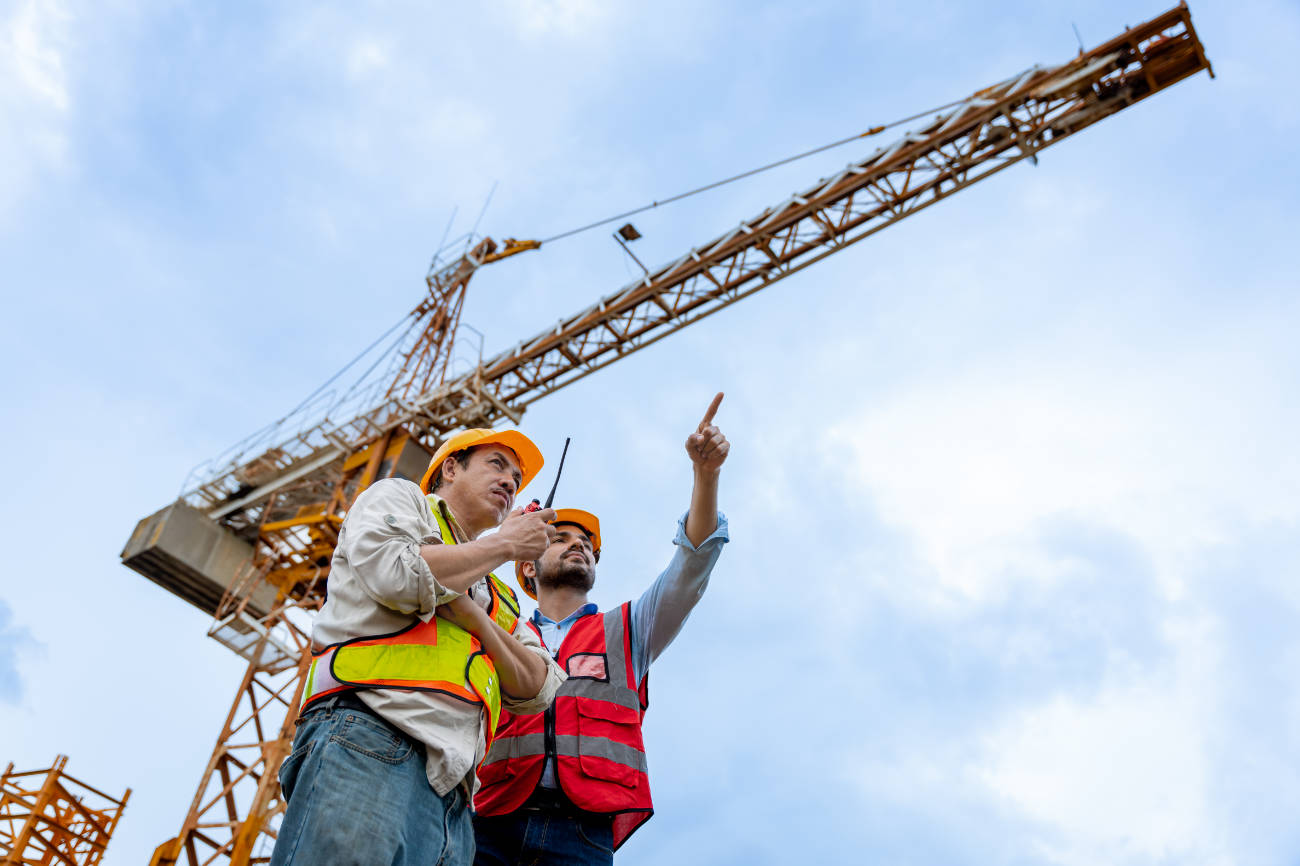 Two men are standing on a construction site, pointing at a crane